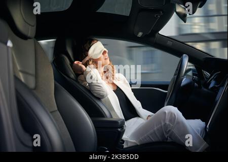 Woman in sleeping mask taking nap in electric car with vehicle autopilot system. Stylish businesswoman sleeping in electro car while using driver-assistance system. Stock Photo