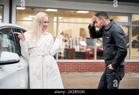 Happy blonde woman showing keys to newly bought white car to her surprised boyfriend outdoors on background of automobil dealership. Anxious man covering his forehead with hand, looking to new buy. Stock Photo