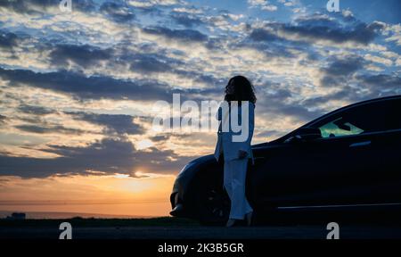 Beautiful sunset. Silhouette of girl standing on road, enjoying incredible sunset over horizon at cloudy sky. Luxury black car parked to her right. Stock Photo