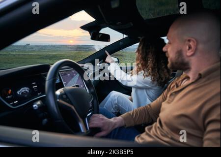 Stylish young woman sitting on passenger seat and pointing finger at sunset while man driving electric car. Female traveler sitting in automobile next to driver and gesturing towards beautiful sky. Stock Photo