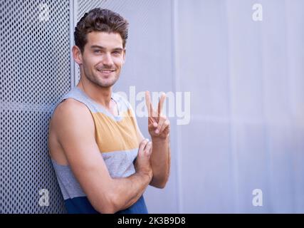 You catch my drift. Portrait of a young man leaning against a wall and flashing a peace sign to the camera. Stock Photo