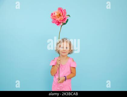 Portrait of smiling little girl hold raising hand with huge artificial flower pink peony over head on blue background. Stock Photo