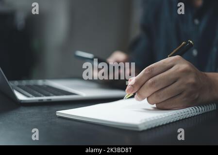 Man hand is writing on a notepad with a pen and a smart phone on desk. Stock Photo