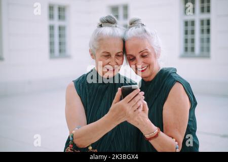 Senior women twins outdoors in city checking smartphone. Stock Photo