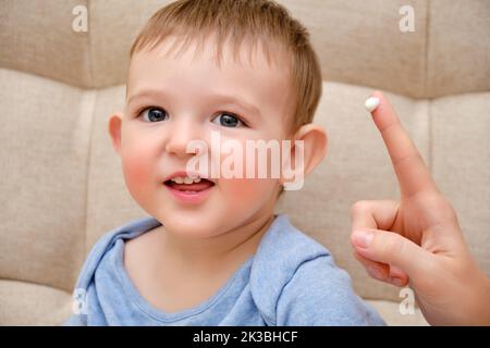 Mother smears allergy cream on face of toddler baby, sofa background in home living room. Close-up portrait of a cute baby and woman hand with cream K Stock Photo