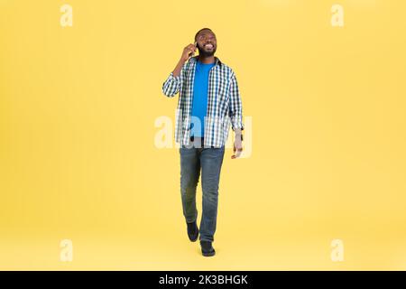 African Guy Talking On Phone Communicating Walking Over Yellow Background Stock Photo
