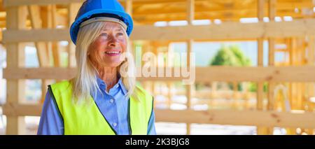 Portrait of elderly woman with protective helmet and yellow vest working at sustainable wooden eco building. Stock Photo