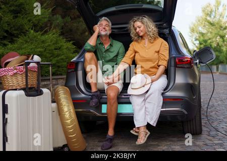 Middle-aged couple sitting in trunk while waiting for charging car before travelling on summer holiday. Stock Photo
