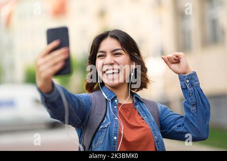 Overjoyed arab female student holding smartphone and celebrating success outdoors Stock Photo
