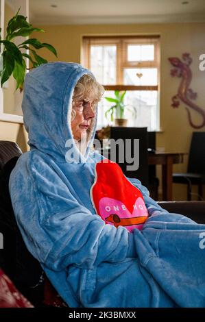 Woman wearing a fleece with hood or hoodie and holding a hot water bottle to keep warm at home as energy costs go up in the UK (posed by model) Stock Photo