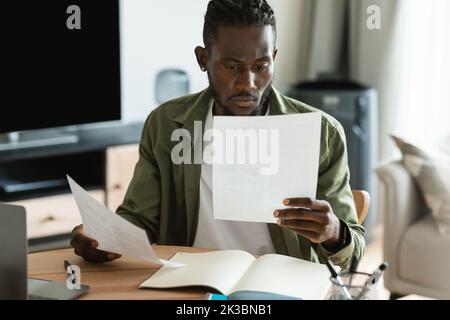 Concentrated african american man checking documents, calculating bills and taxes using laptop in room interior Stock Photo