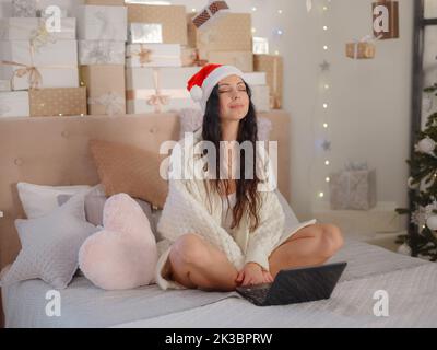 Beautiful woman sitting on bed working with laptop at home around christmas lights relax and smiling with eyes closed doing meditation. Yoga concept. Stock Photo