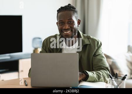 Cheerful african american guy programmer sitting at workdesk at home, male freelancer sitting in front of computer Stock Photo