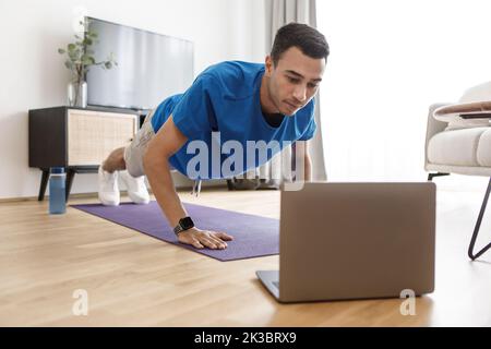 Handsome arab guy in sportswear doing push-ups, having online fitness class via laptop from home, copy space Stock Photo