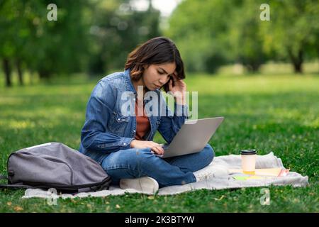 Upset Arab Female Student Using Laptop While Sitting On Plaid In Park Stock Photo