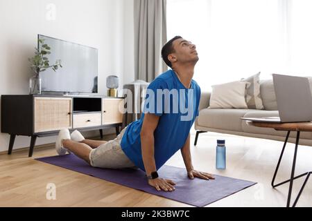 Young arab man practicing pilates with pc, watching online tutorials and standing in cobra pose Stock Photo