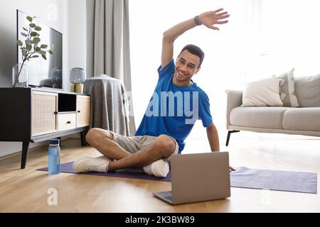 Positive arab man having fitness class online at home, using laptop, watching sport videos on Internet and stretching Stock Photo