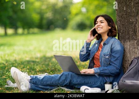 Smiling Young Arab Female Student Talking On Cellphone And Using Laptop Outdoors Stock Photo