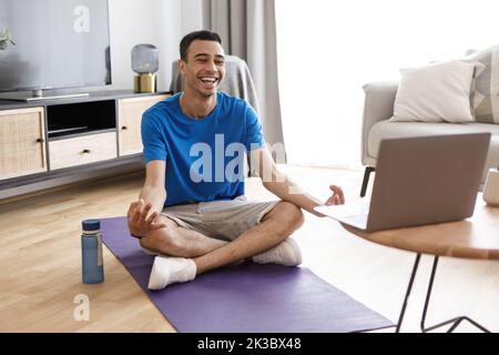 Positive calm arab man in sportswear sitting in lotus pose, having online yoga class, meditating near laptop at home Stock Photo