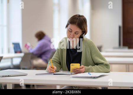 Concentrated mature female student using smartphone for exam preparation, taking second degree Stock Photo