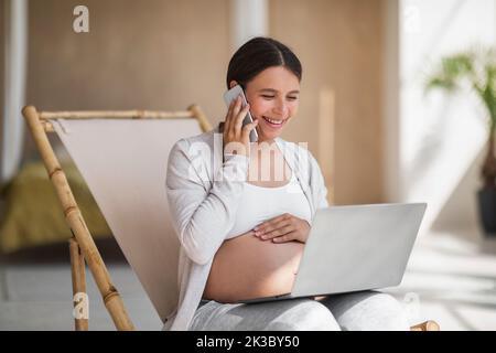 Remote Work Concept. Happy Pregnant Lady Using Laptop And Talking On Cellphone Stock Photo