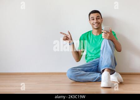 Happy arab guy pointing finger aside at copy space and using smartphone, sitting on floor against white studio wall Stock Photo