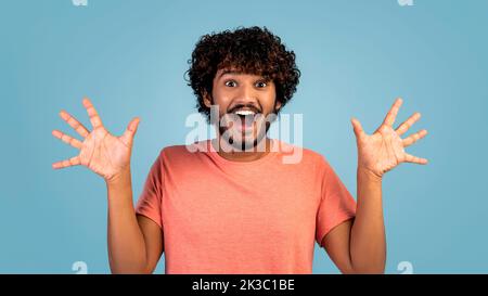 Amazed hindu guy raising hands up over blue background Stock Photo