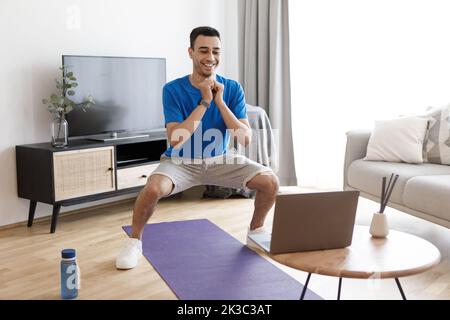 Young arab man doing deep squats, looking at laptop during online training, exercising at home, free space Stock Photo