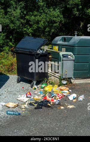 Overflowing trash bin with scattered litter beside recycling containers in a public area Stock Photo