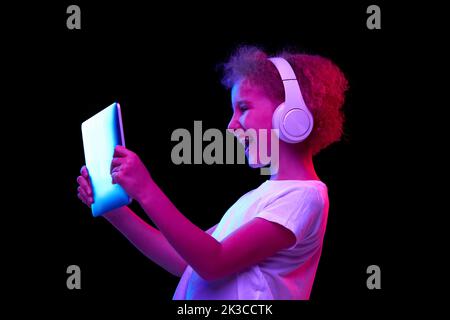 Emotional curly school age girl in casual tee using digital tablet isolated on dark background in neon light. Stock Photo