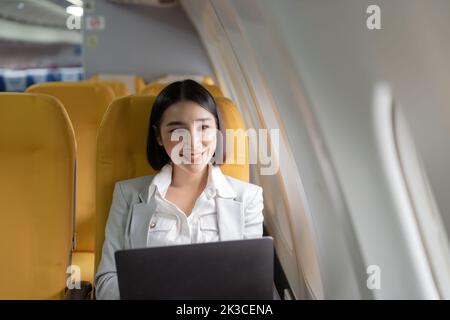 Traveling and technology. Flying at first class. Pretty young businees asian woman working on laptop computer while sitting in airplane. Stock Photo