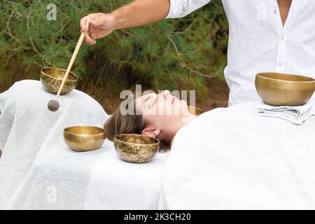 Young woman laying on a massage bed with tibetan singing bowl outdoor. A master of sound massage therapy doing a Buddhist healing practices, male hand Stock Photo