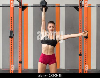 Muscular female athlete in sports and bra stretching out arm and doing push press exercise with heavy dumbbell during functional workout in gym Stock Photo