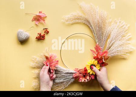 Hands making dried floral wreath from dry pampas grass and Autumn leaves. Flat lay on white yellow table, sunlight with long shadows. Stock Photo