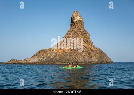 Two kayakers in front of Faraglione Grande, one of the Faraglioni or Isole dei Ciclopi, a group of volcanic basalt sea stacks off Aci Trezza, Sicily Stock Photo