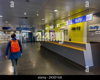 Basel, Switzerland - Sep 22, 2022: Side view of young handsome solo traveler with red backpack passing near rent-a-car rental company desk in international airport Hertz Thrifty Stock Photo