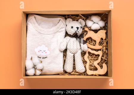 Gift basket with gender neutral baby garment and accessories. Care box of organic newborn cotton clothes, teddy, cotton in box on orange background Flat lay Top view. Stock Photo