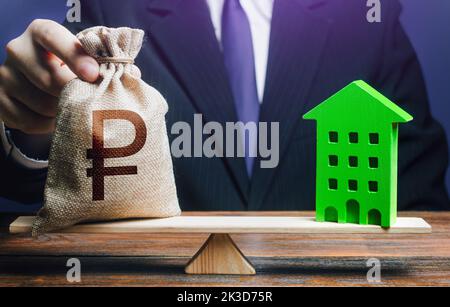 Businessman puts russian ruble money bag on scales and green house. Investment in renovation. Profitability housing of eco technologies. Reduced CO2 e Stock Photo