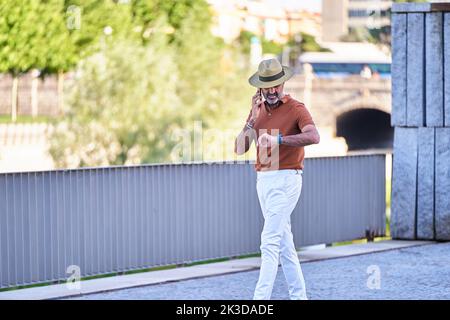 Man in hat looking at his watch while talking on the phone walking outdoors. Technology and communication concept. Stock Photo