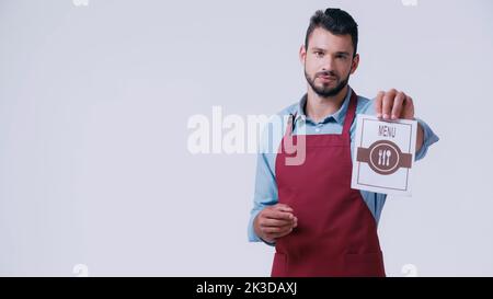 young bearded waiter in apron showing menu and looking at camera isolated on grey,stock image Stock Photo