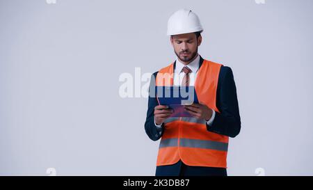 thoughtful engineer in helmet and safety vest looking at clipboard isolated on grey,stock image Stock Photo