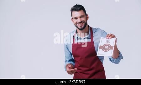 happy young waiter in apron showing menu isolated on grey,stock image Stock Photo