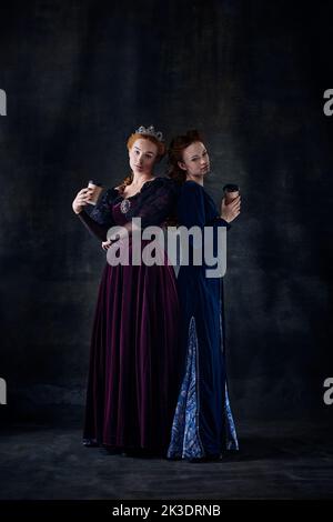 Portrait of two beautiful women in image of queens isolated over dark background. Coffee break Stock Photo