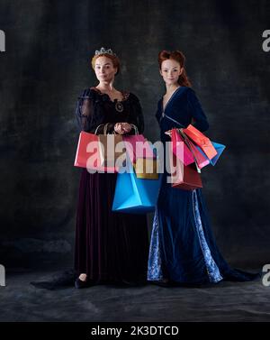 Portrait of two beautiful women in image of queens with many shopping bags isolated over dark background Stock Photo