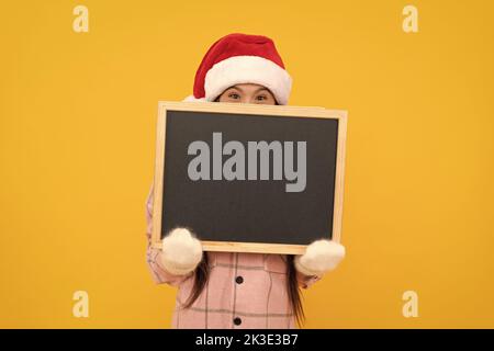 kid in santa hat behind school blackboard with copy space on yellow background, advertising. Stock Photo