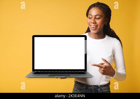 Positive smiling female freelancer holding and showing laptop with empty screen, satisfied multiracial woman advertising new computer app isolated on yellow background, mock-up Stock Photo