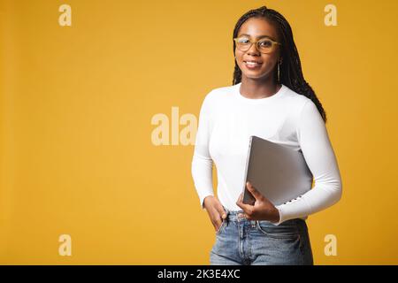 Young African American woman wearing stylish eyeglasses standing isolated on yellow, carrying laptop computer, smiling, female office employee, student Stock Photo
