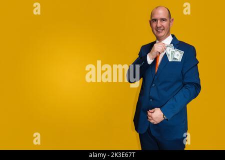 A bald Caucasian adult male wearing a blue suit, white shirt and orange tie pulling one dollar bills from his jacket pocket. The background is yellow. Stock Photo