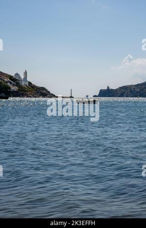 View of a little fishing boat with a Greek flag, a beautiful church and the lighthouse in the background at the port of Mylopotas in Ios Greece Stock Photo