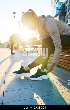 Side view of young blond sportswoman bending over leg bent in knee while sitting on bench in urban environment during break Stock Photo
